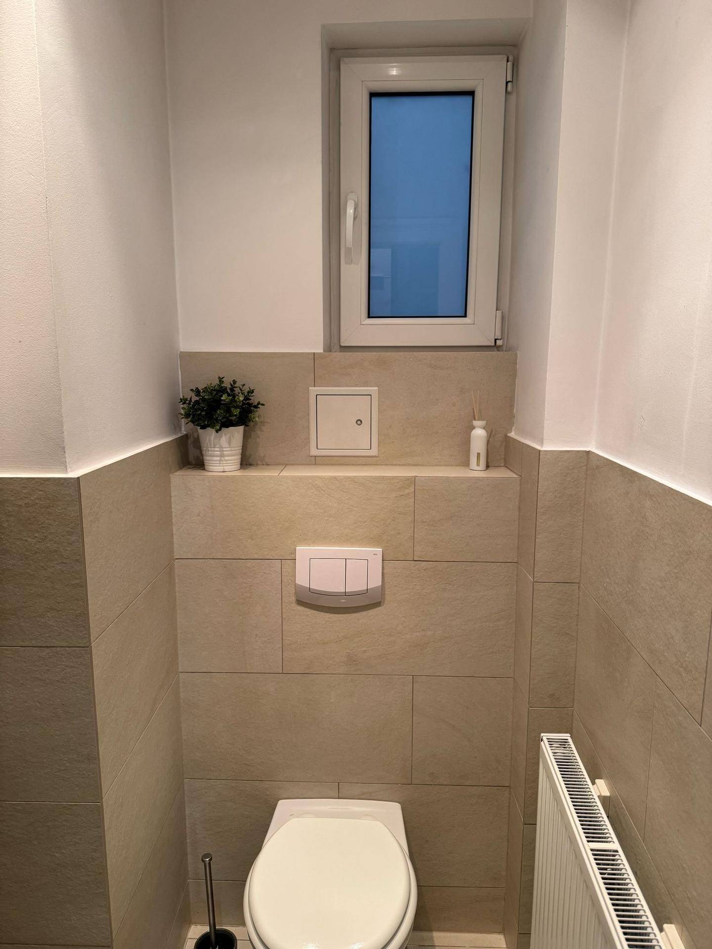 Kleines Gäste-WC mit beigefarbenen Fliesen, wandmontierter Toilette und einem Fenster für Tageslicht.