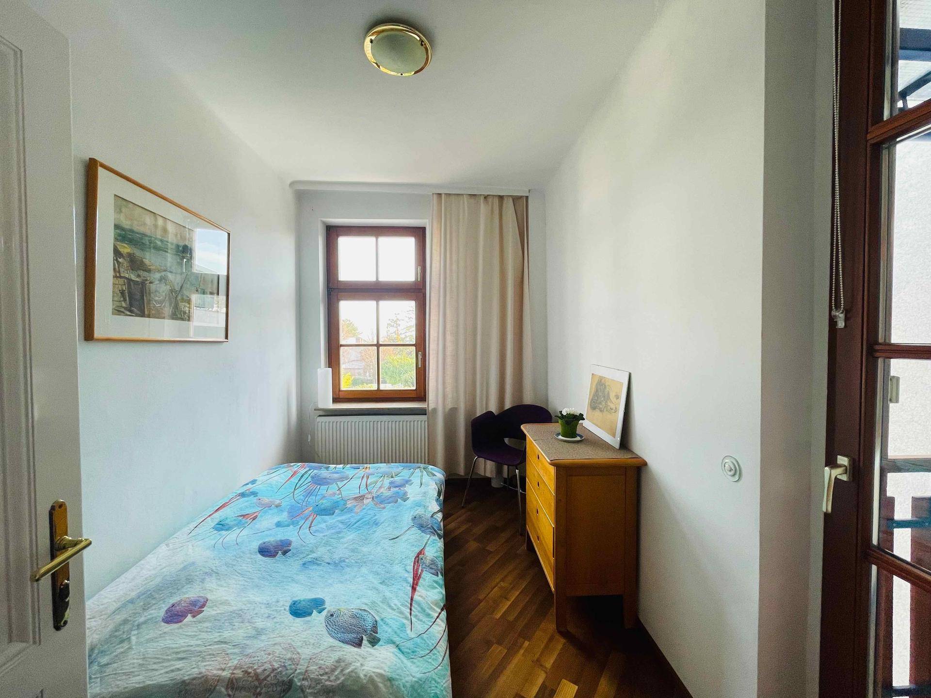 Gemütliches Schlafzimmer mit Einzelbett, Holzkommode, Fenster und Zugang zu einem Balkon.