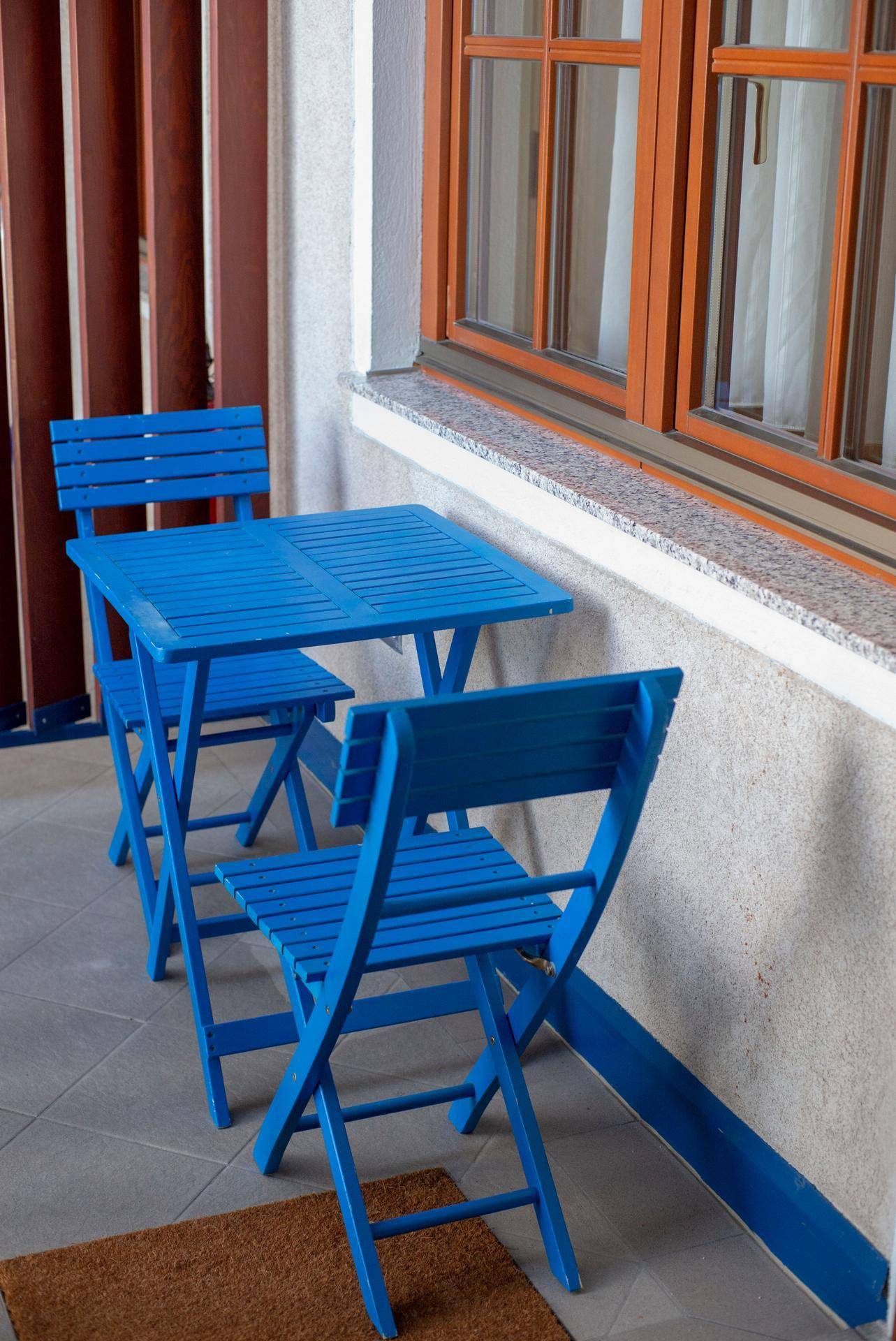 Gemütlicher Außenbereich mit blauem Tisch und Stühlen auf gefliestem Boden neben einem Fenster.