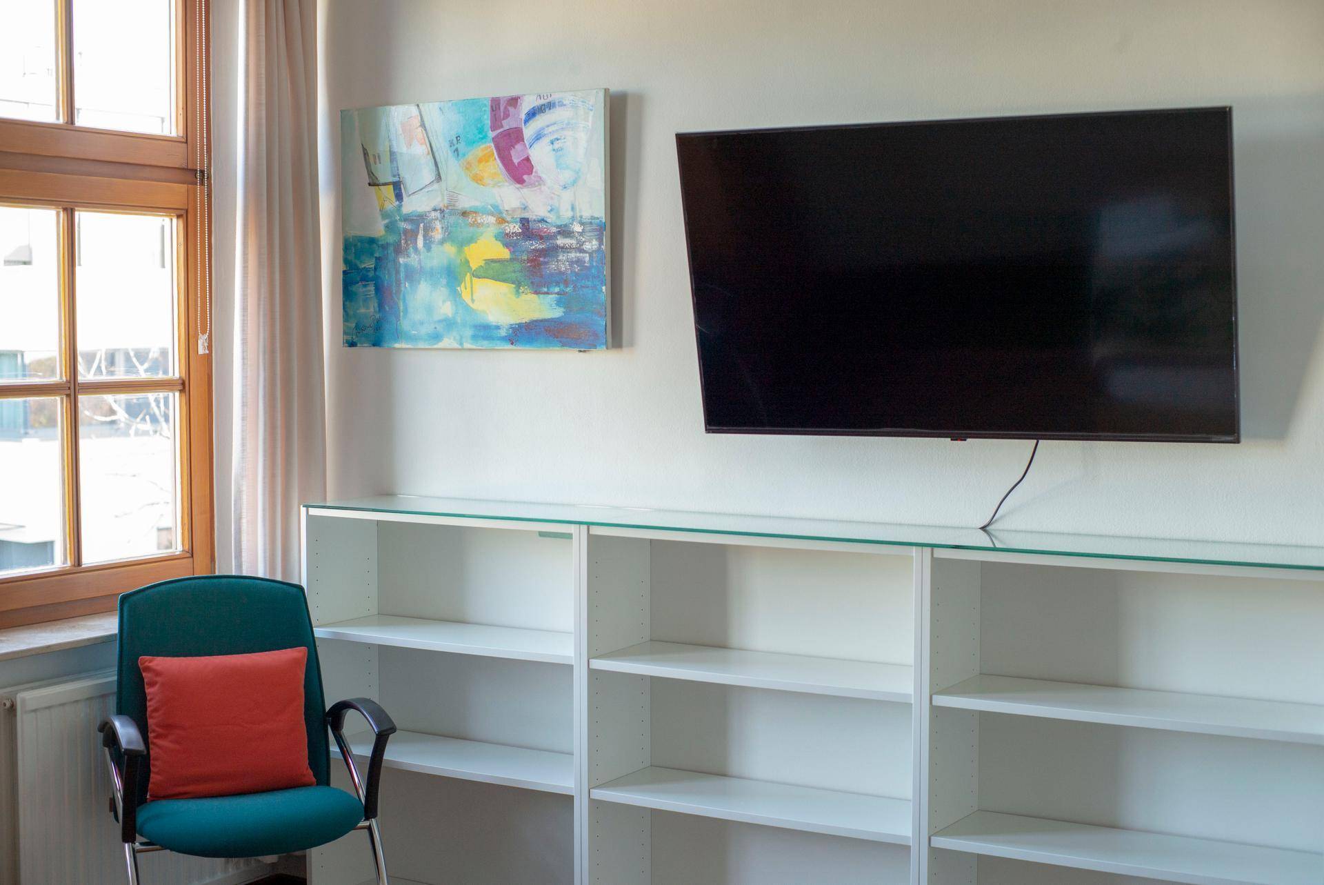 Heller Raum mit Wandfernseher, weißen Regalen und einem Fenster, ideal als Wohn- oder Arbeitsbereich.