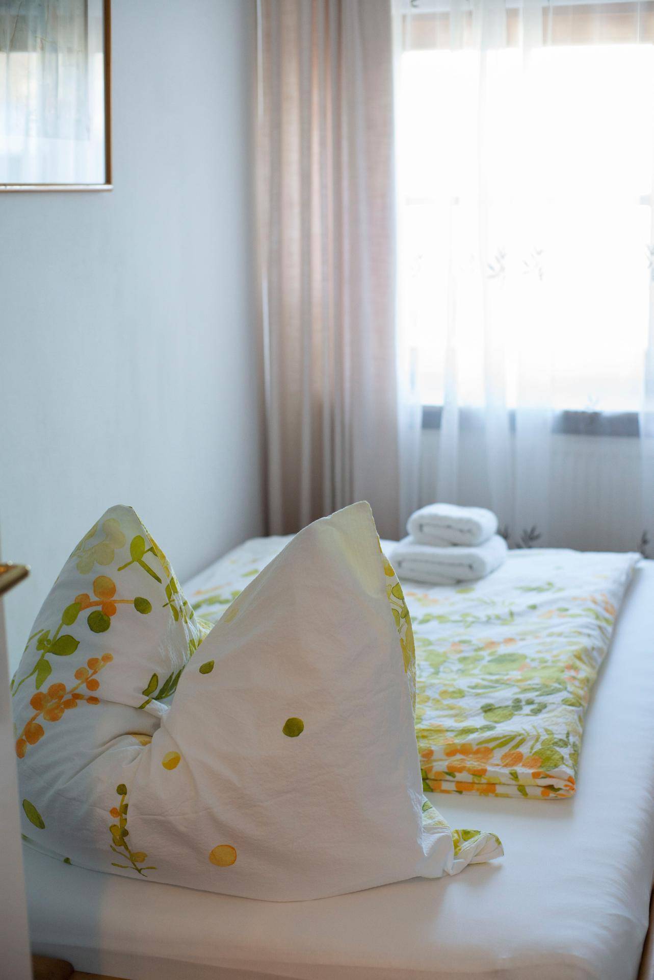Nahaufnahme eines Bettes mit floraler Bettwäsche, weißen Handtüchern und hellem Fenster mit Vorhängen.