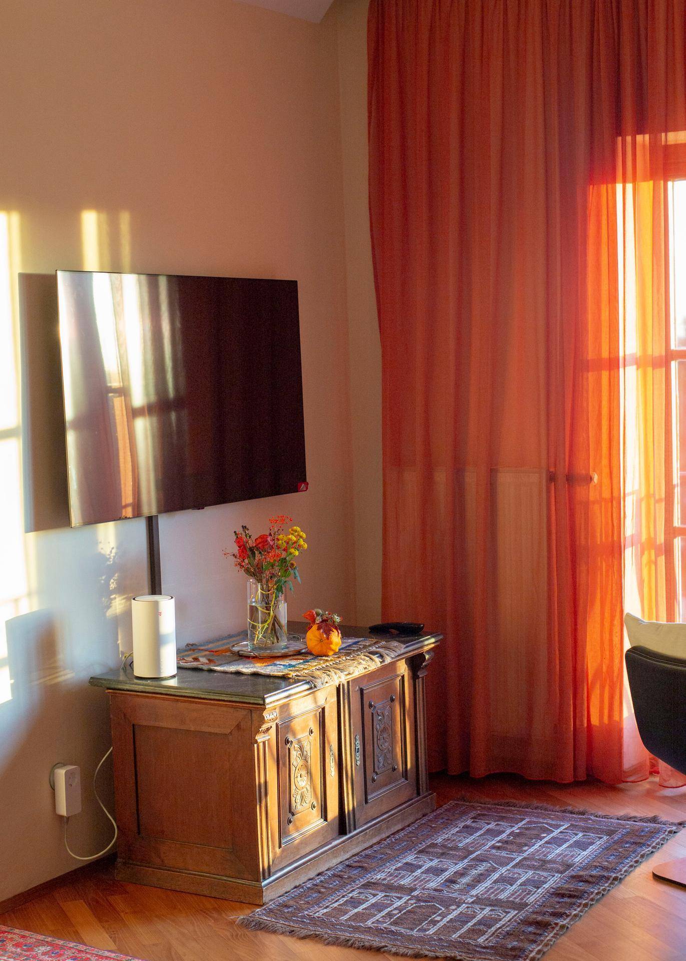 Wohnzimmer mit Wandfernseher, antiker Kommode und orangefarbenen Vorhängen am Fenster.