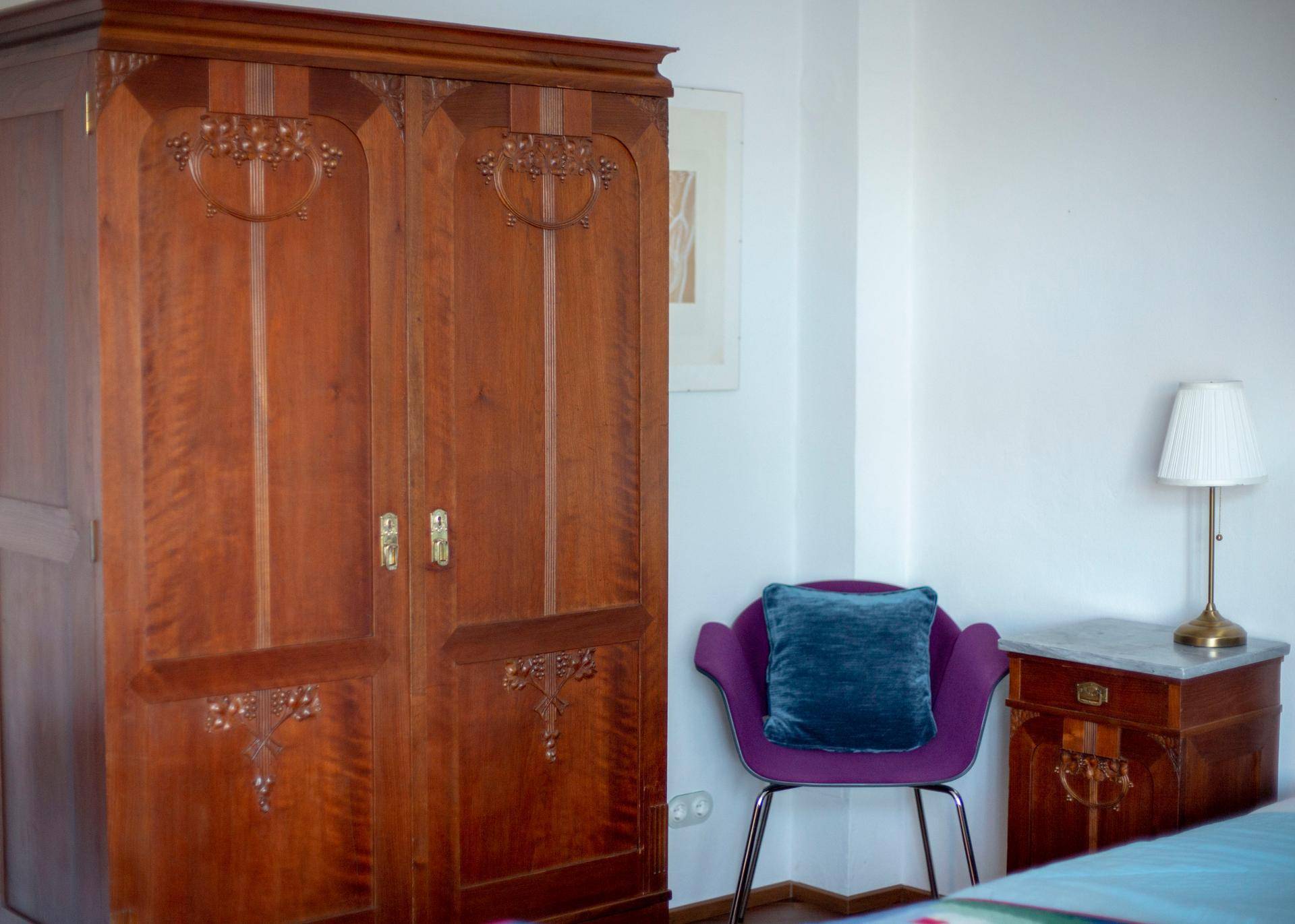 Antiker Holzschrank mit geschnitzten Details und lila Sessel im Schlafzimmer.