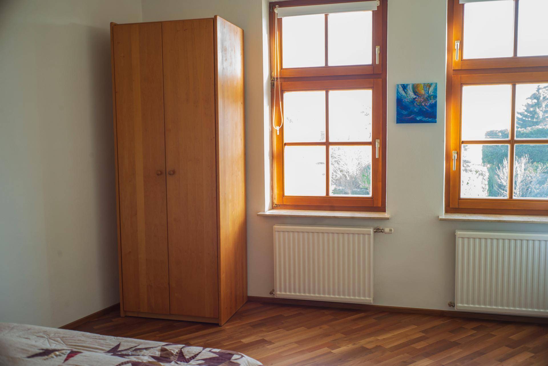 Helles Schlafzimmer mit Holzschrank und zwei Fenstern, die viel Tageslicht hereinlassen.
