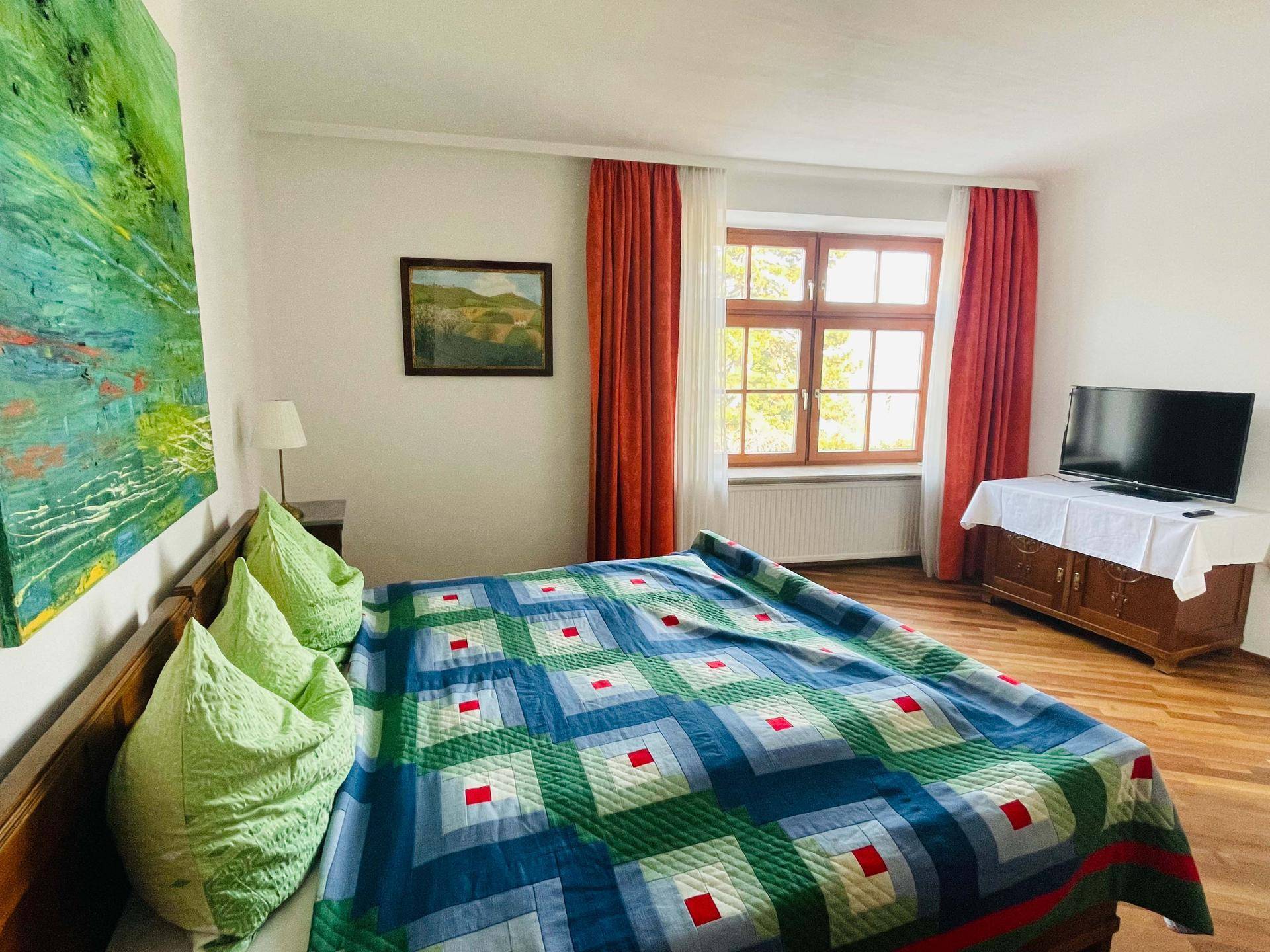 Helles Schlafzimmer mit Doppelbett, Fenster und Fernseher auf einem antiken Schrank.