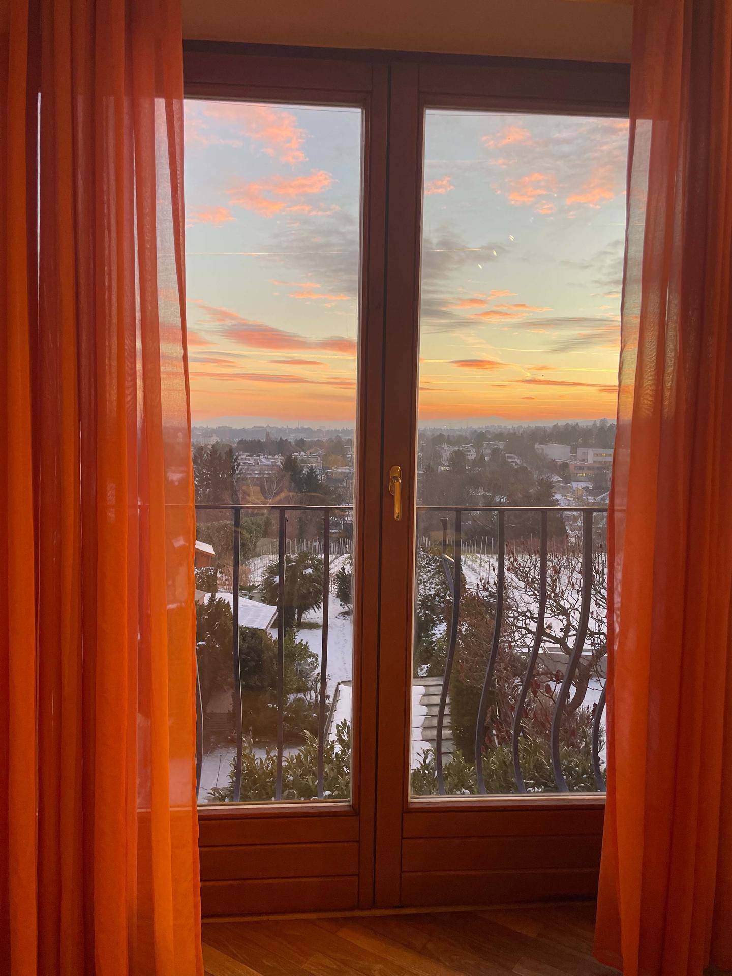 Sonnenuntergang über der Stadt, gesehen durch ein großes Fenster mit orangefarbenen Vorhängen.