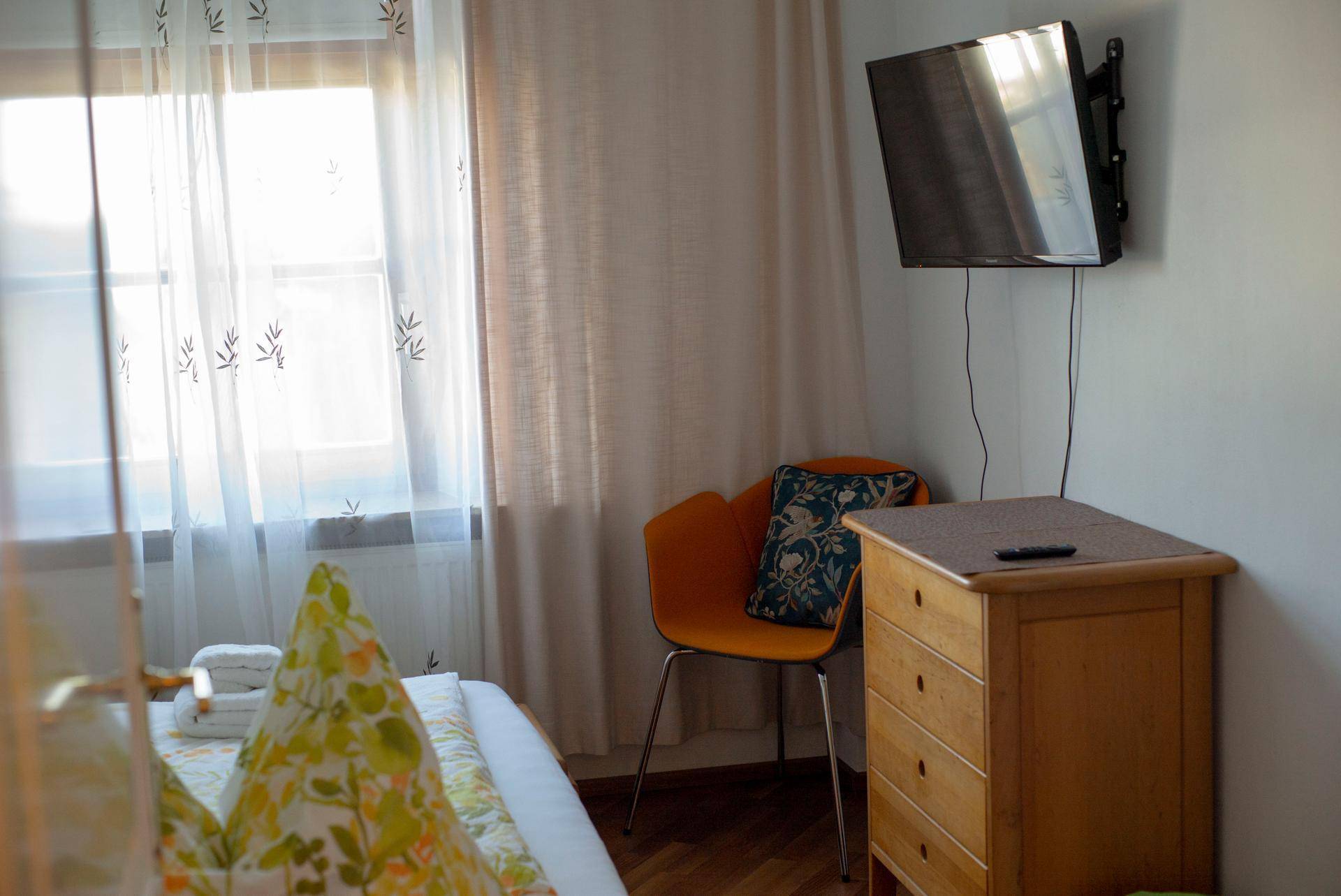 Schlafzimmerecke mit Bett, Holzkommode, Wandfernseher und einem hellen Fenster mit Vorhängen.