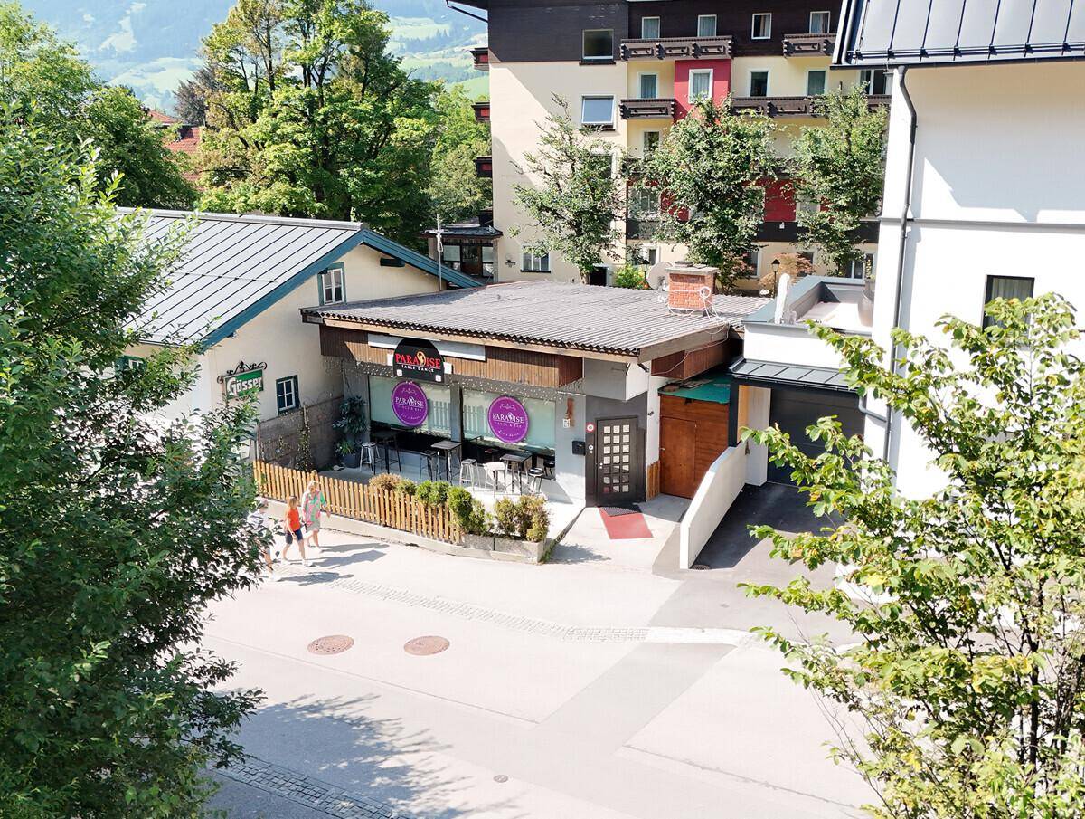 Außenansicht eines Geschäftsgebäudes mit Terrasse und angrenzendem Wohnhaus in bergiger Umgebung.