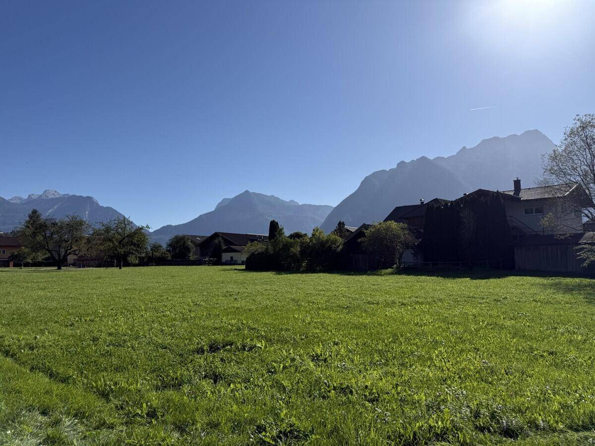 Weitläufige grüne Wiese unter strahlend blauem Himmel mit Häusern und Bergen im Hintergrund, sonnendurchflutet.