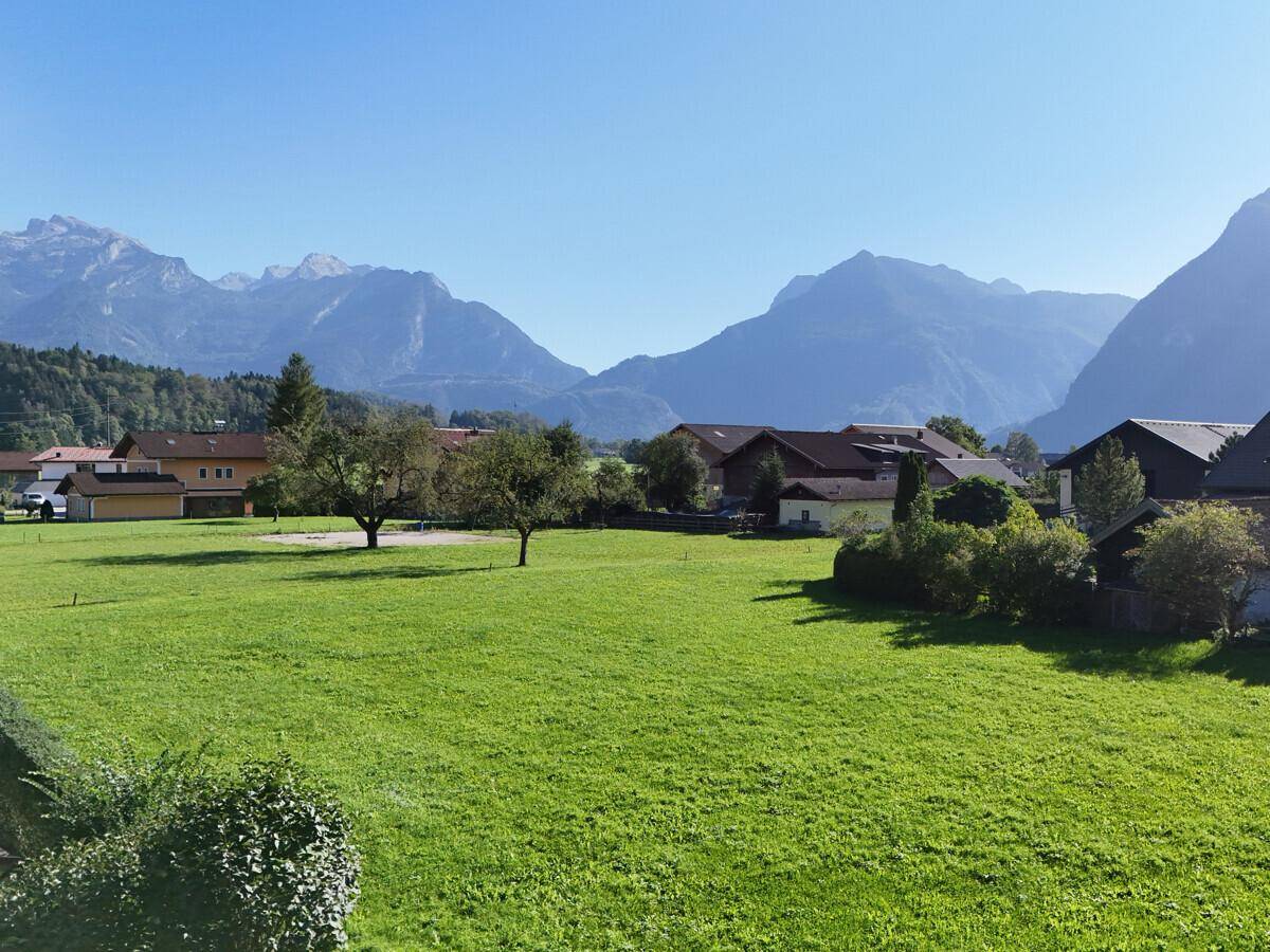 Weitläufiges grünes Feld mit vereinzelten Häusern und einer klaren Sicht auf die umliegende Berglandschaft.