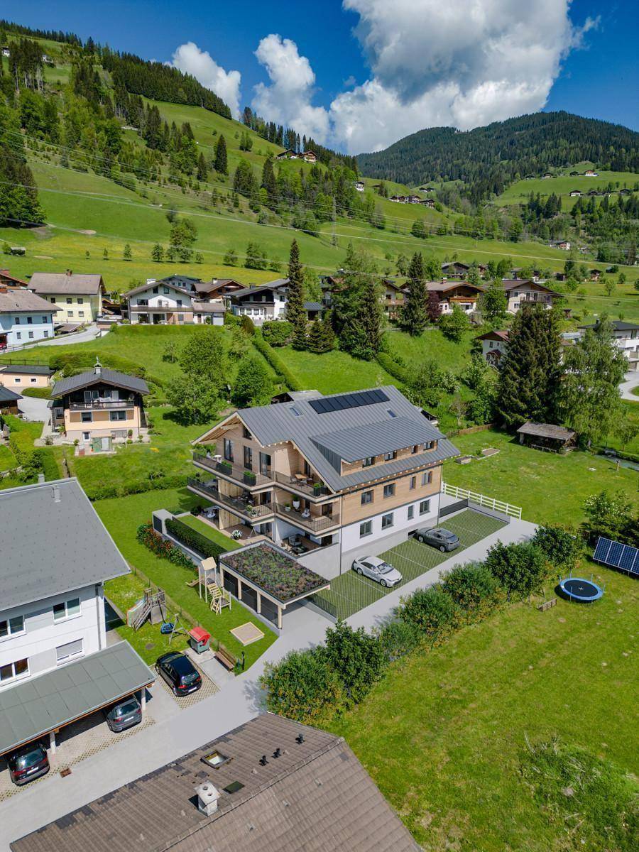 Luftaufnahme des modernen Apartmenthauses in einer grünen Berglandschaft mit Parkplätzen und Spielplatz.