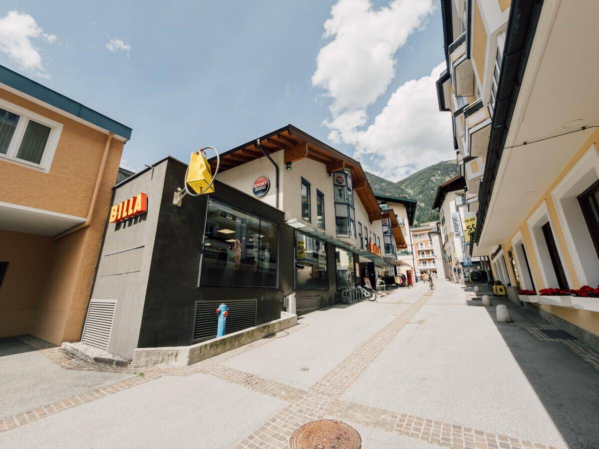 Straßenansicht des Supermarktes und angrenzender Gebäude in einer sonnigen Berglandschaft.