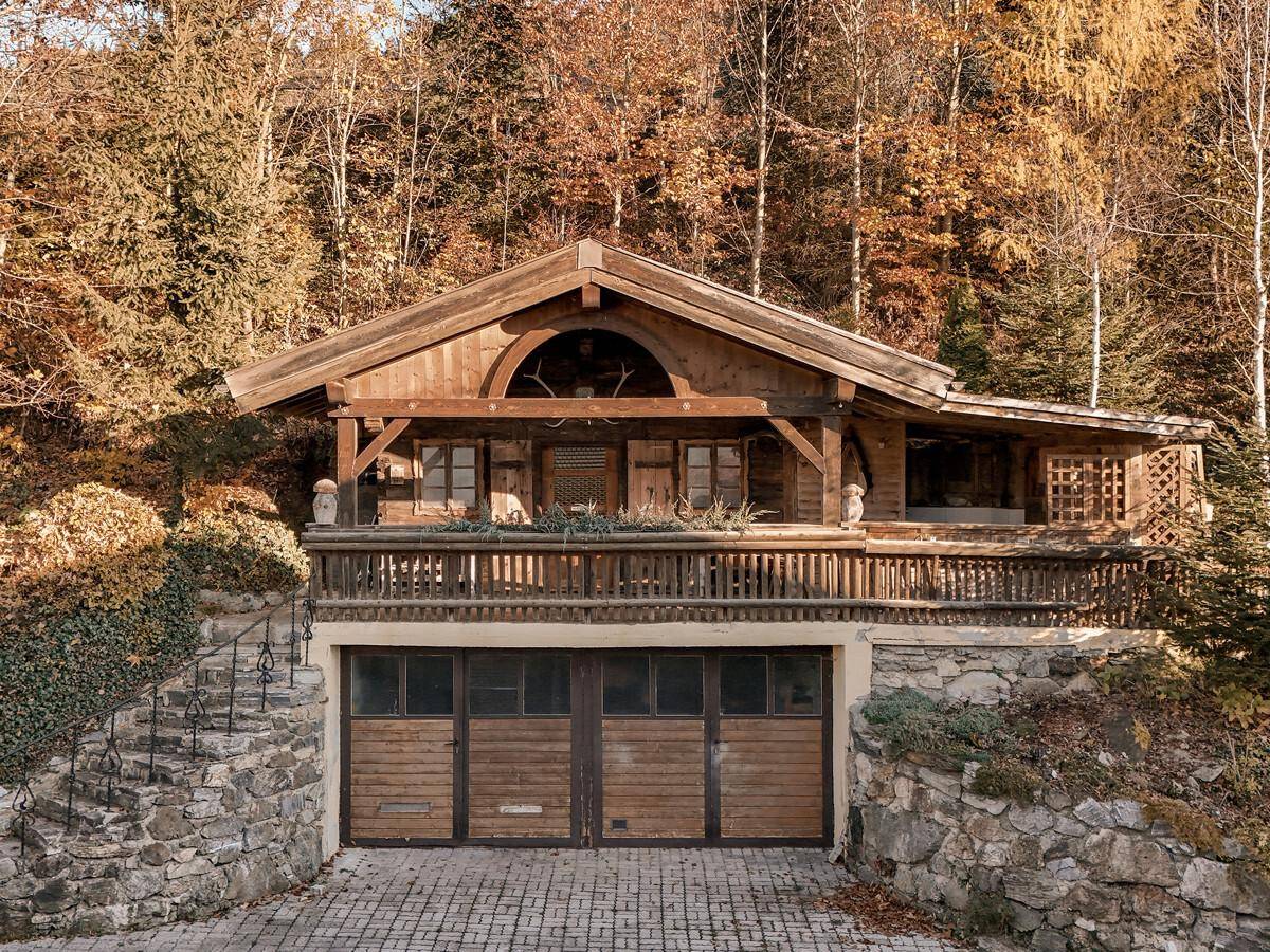 Traditionelles Holzchalet mit Steinfundament und integrierter Garage, umgeben von herbstlichen Bäumen.