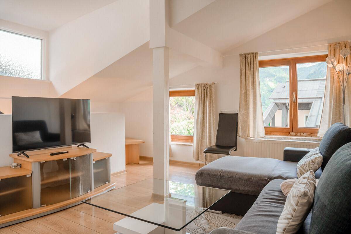 Geräumiges Wohnzimmer mit Sofa, Fernseher und großen Fenstern, die viel Tageslicht hereinlassen.