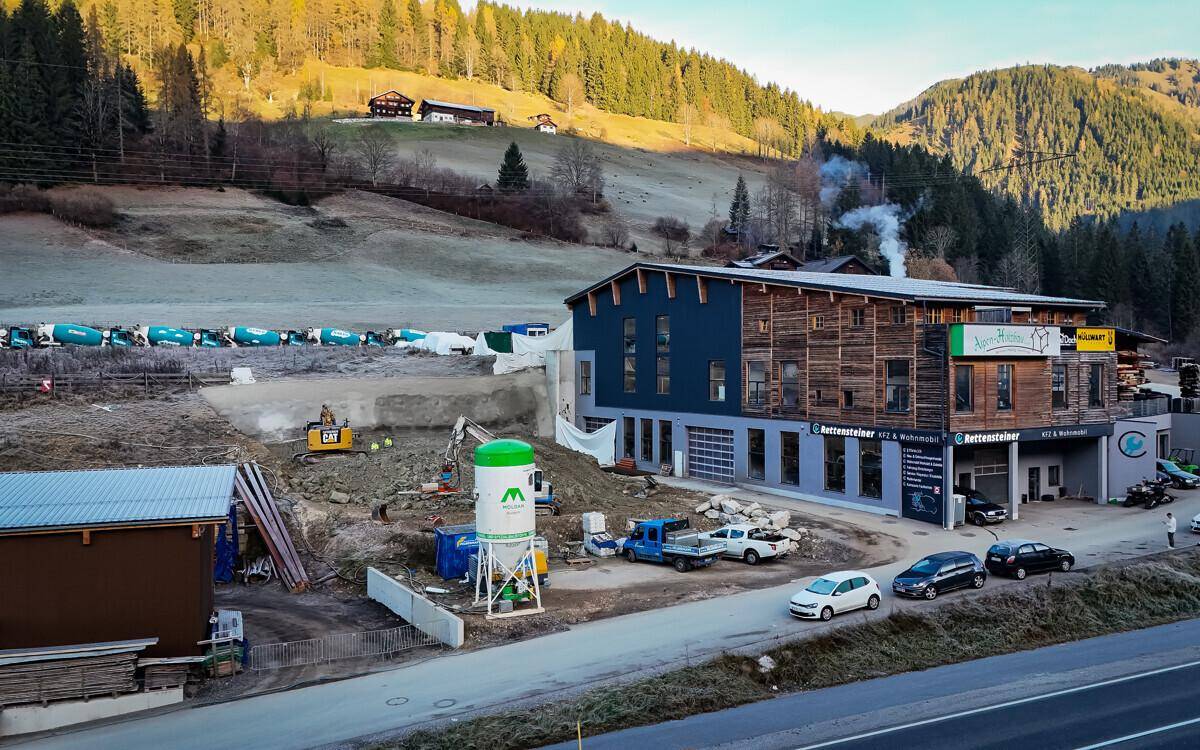 Blick auf ein modernes Gebäude mit Holzfassade und Baustelle in ländlicher Umgebung mit Bergen und grünen Hügeln.