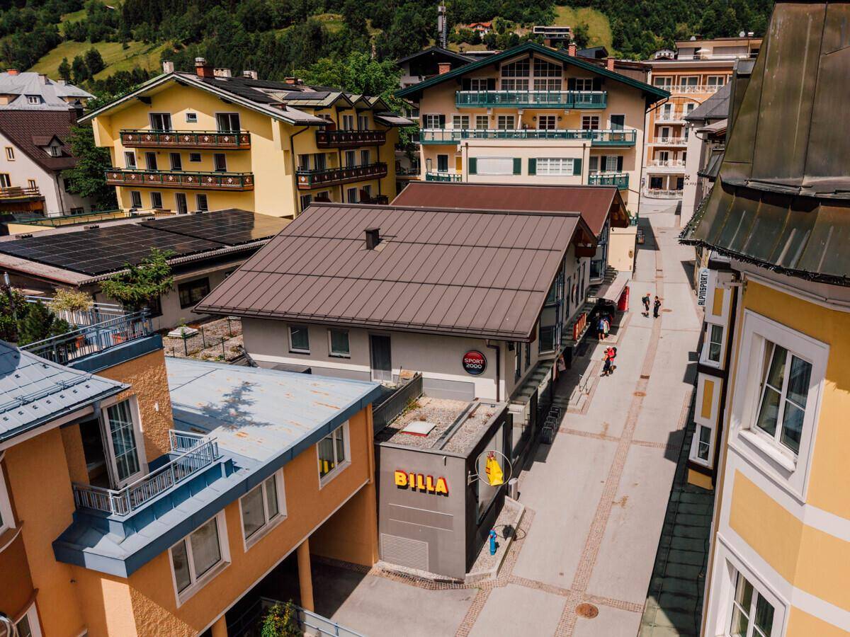 Luftaufnahme einer belebten Straße mit verschiedenen Gebäuden und Supermarkt in alpiner Umgebung.