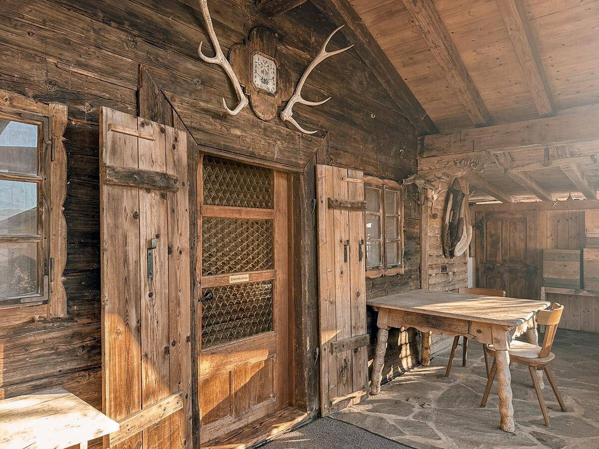 Eingangsbereich eines Holzchalets mit rustikaler Holztür, Fensterläden und traditionellem Hirschgeweih als Dekoration.
