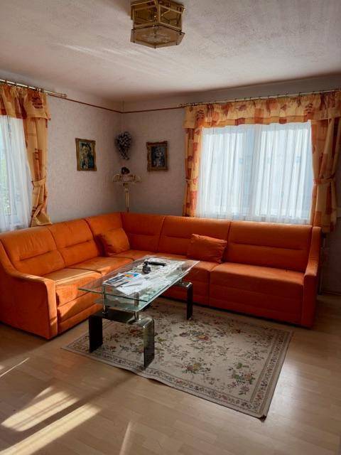 Helles Wohnzimmer mit orangefarbenem Ecksofa, Glastisch und Parkettboden.