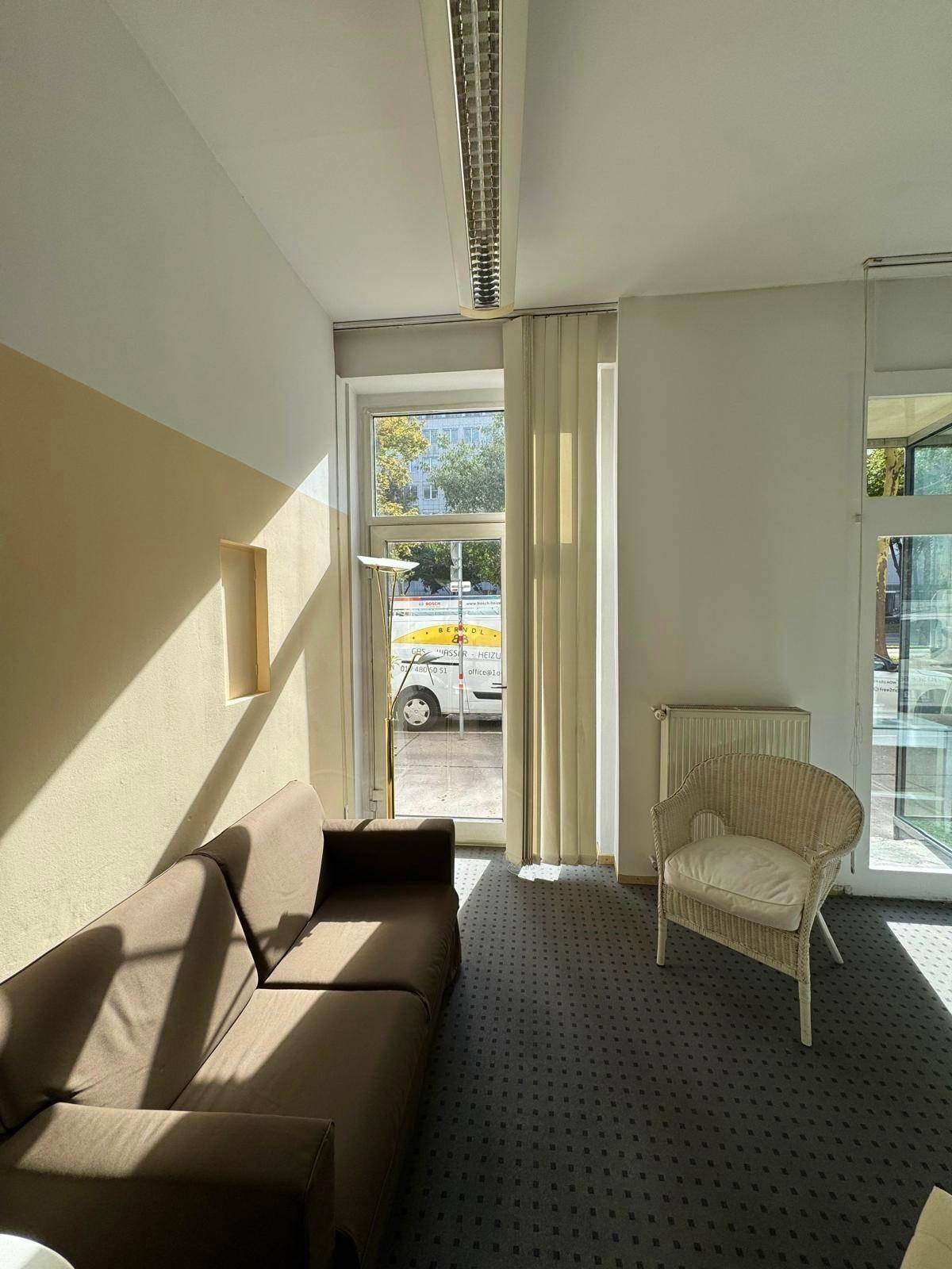 Sonniges Büro mit Sofa, Sessel und großen Fenstern, die viel Tageslicht hereinlassen.