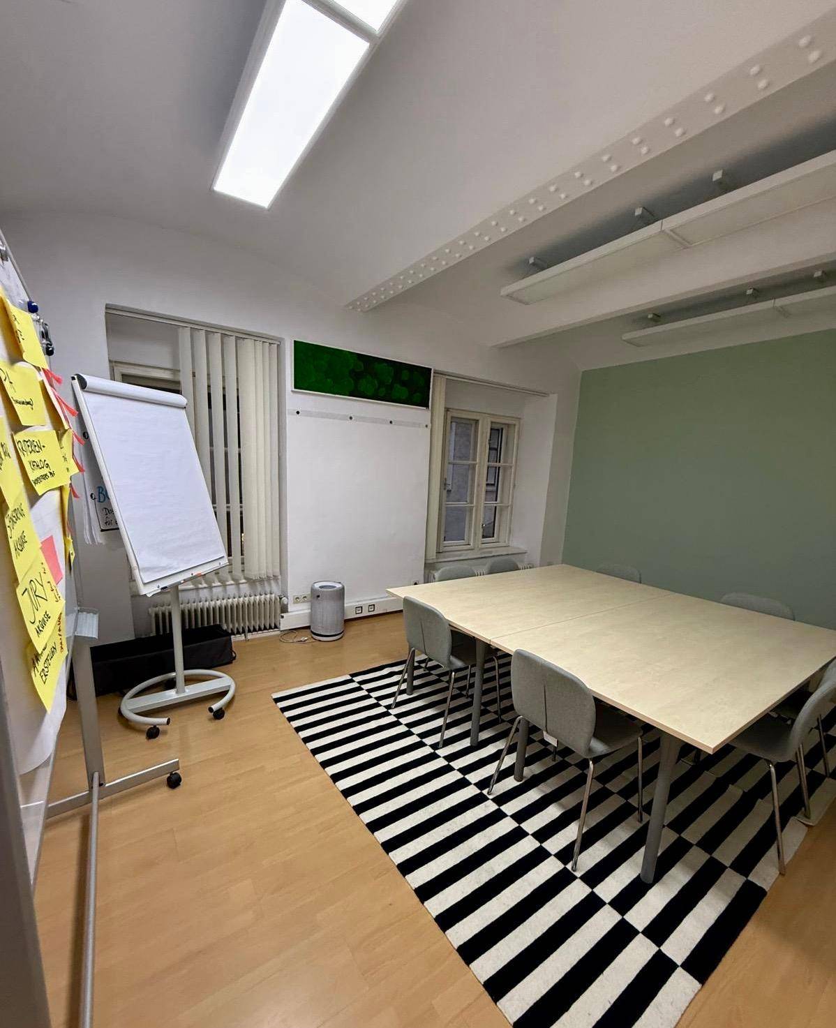 Heller Konferenzraum mit Holzboden, Flipchart und einem schwarz-weiß gemusterten Teppich.
