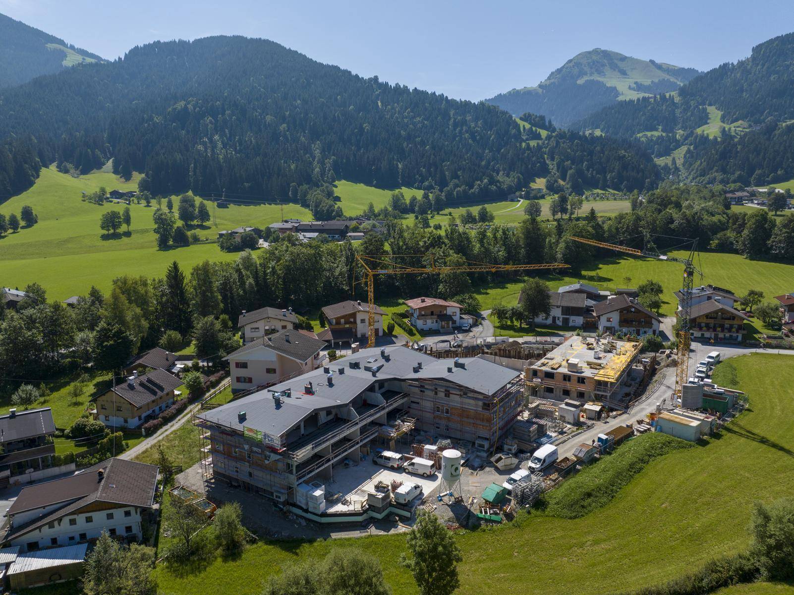 Luftaufnahme einer großen Baustelle für eine Wohnanlage, umgeben von grünen Hügeln und Bergen.