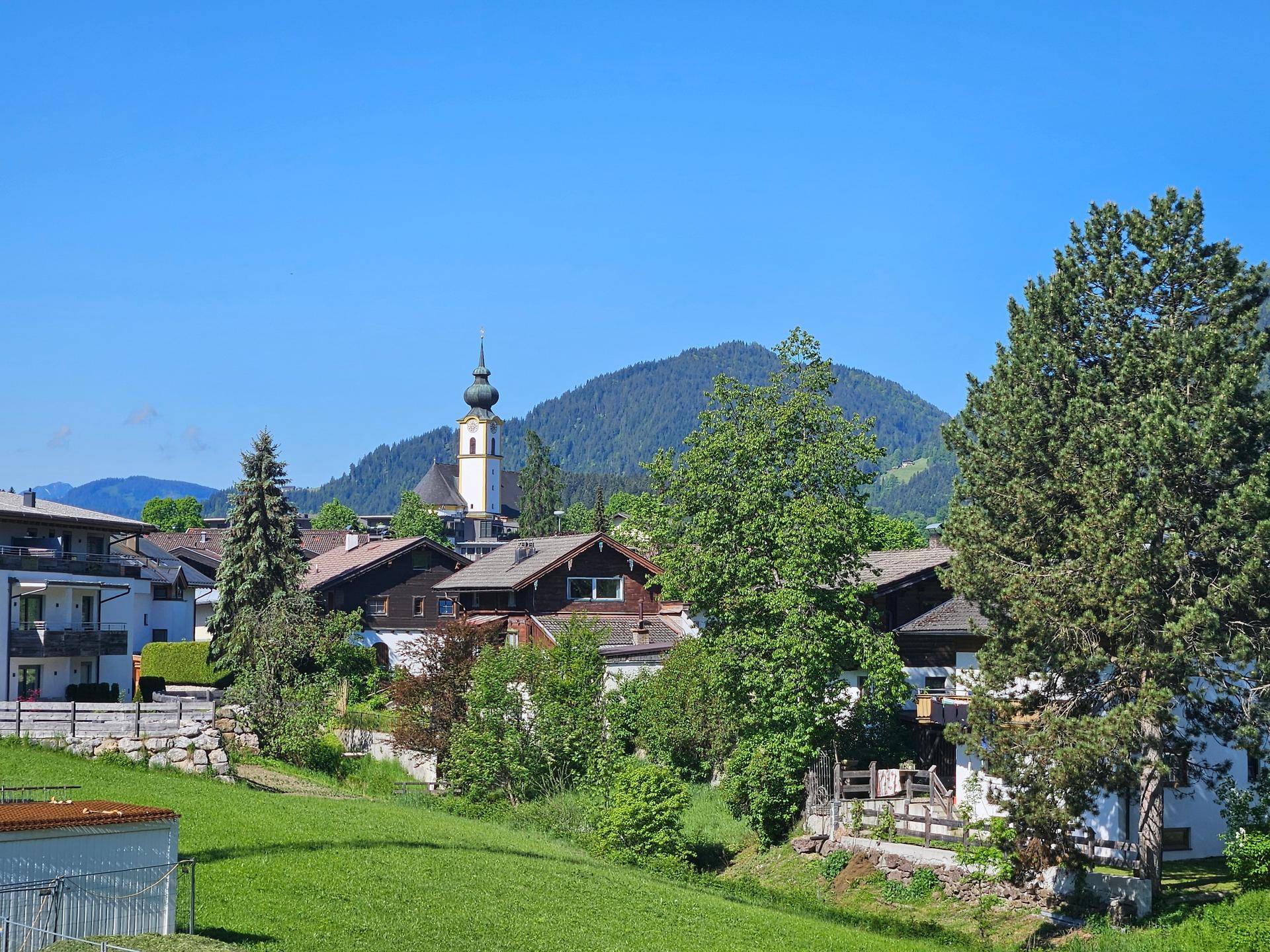 Panoramablick auf ein malerisches Dorf mit Kirche und die umliegende Berglandschaft unter blauem Himmel.