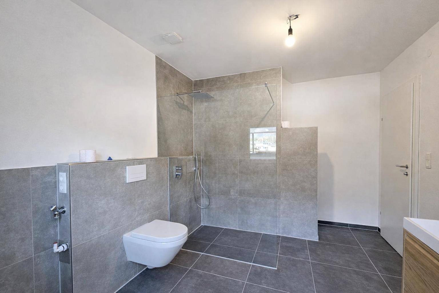 Modernes Badezimmer mit ebenerdiger Dusche, Wand-WC und grauen Fliesen, bereit für die Endmontage des Waschtischs.