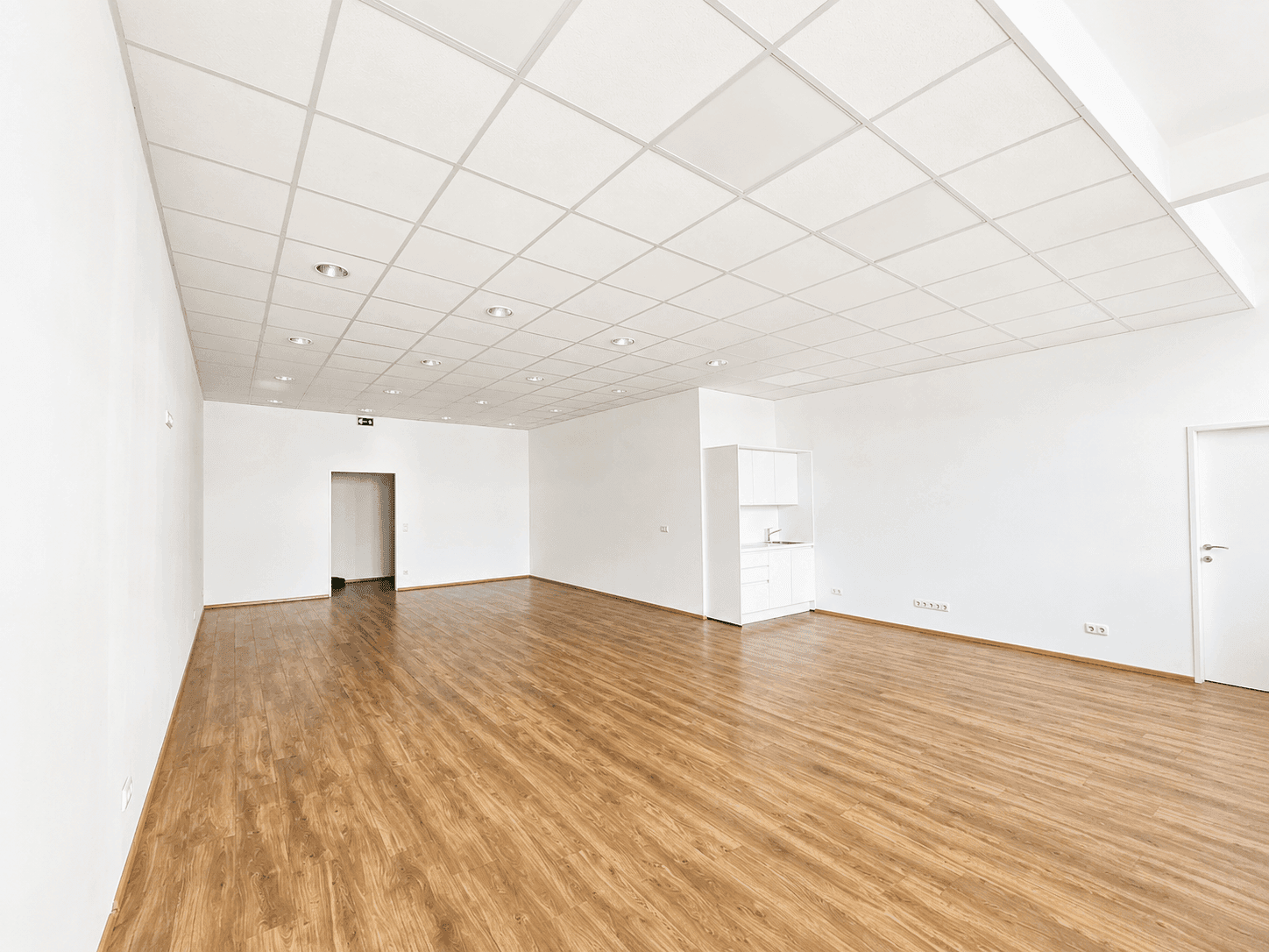 Geräumiger Gewerberaum mit hellem Laminatboden, weißen Wänden und einer kleinen Küchenzeile.