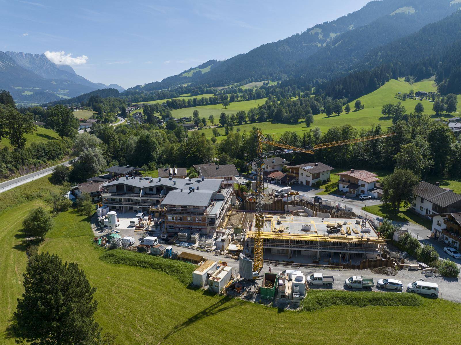 Luftaufnahme einer großen Baustelle für eine Wohnanlage, umgeben von grünen Hügeln und Bergen.