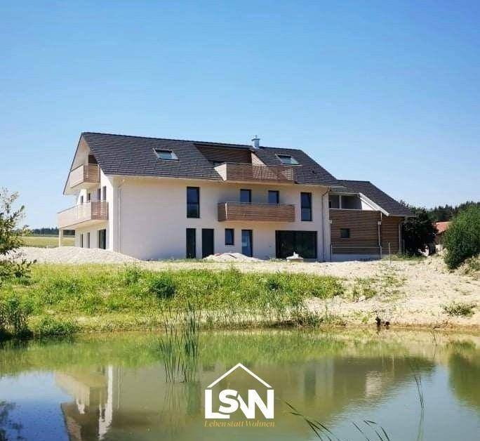 Modernes Wohnhaus mit Balkonen und Teich im Vordergrund, umgeben von grüner Natur und Feldern.