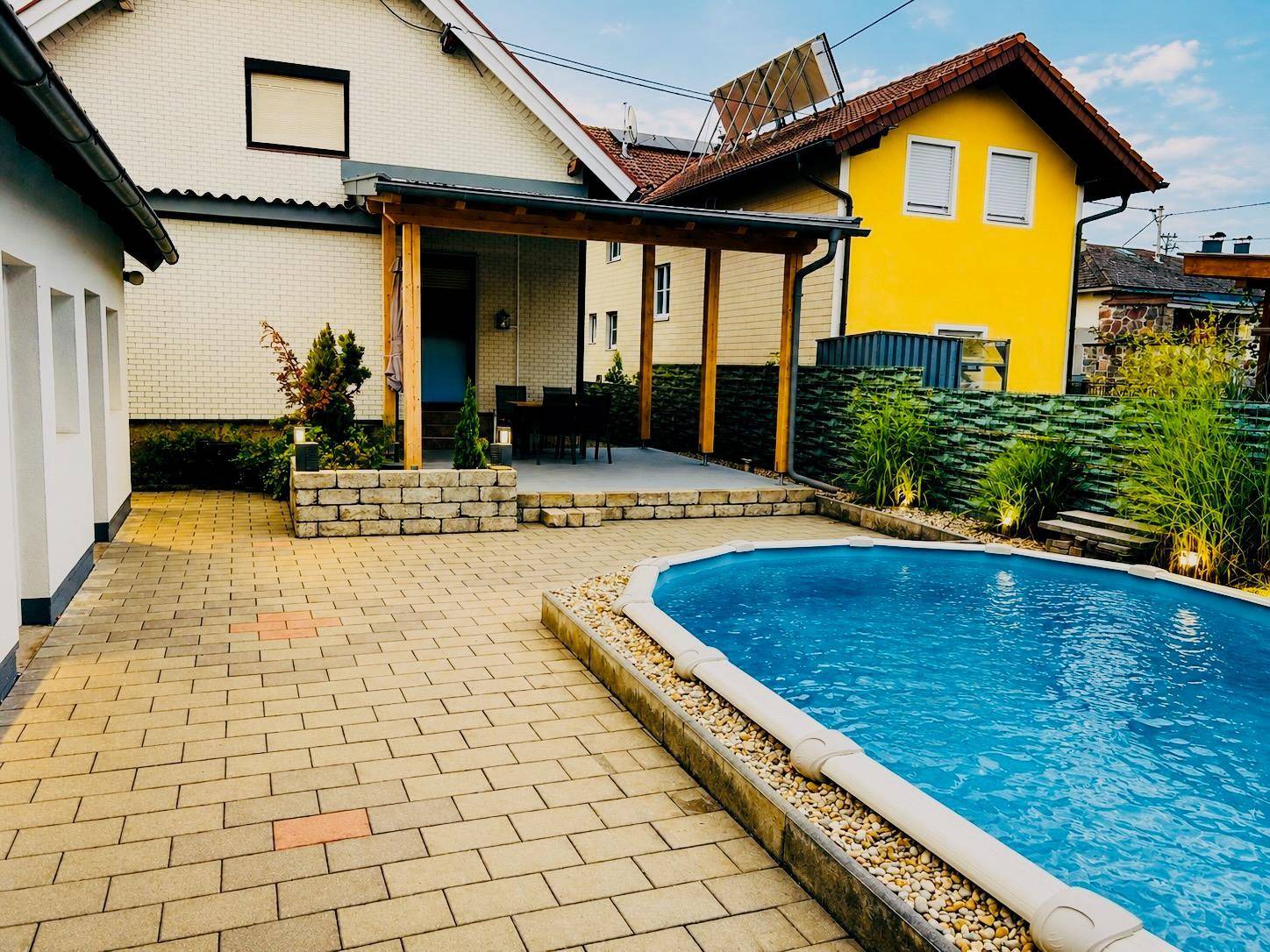 Gepflegter Außenbereich mit Swimmingpool, gepflasterter Fläche und einer überdachten Terrasse im Hintergrund.