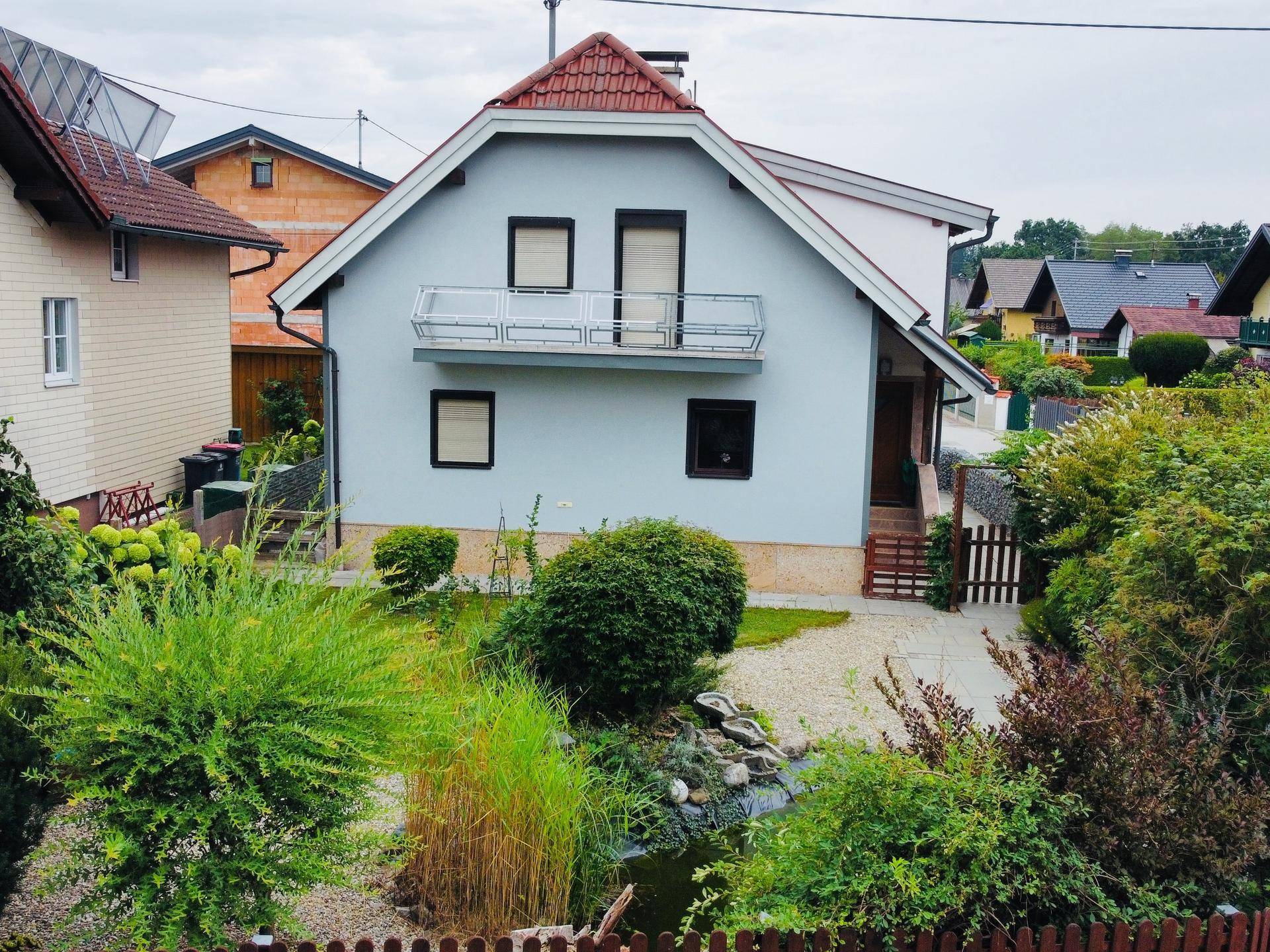 Vorderansicht des Hauses mit hellblauer Fassade, kleinem Teich und üppiger Bepflanzung im Vorgarten.