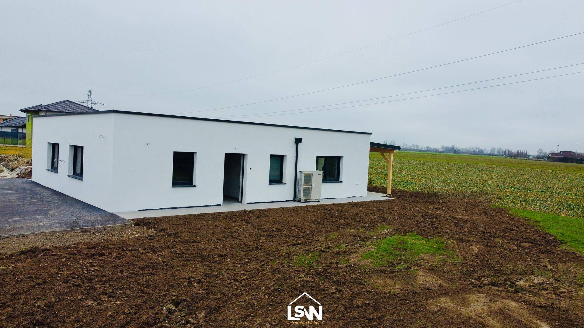 Weißes Einfamilienhaus mit Flachdach und Wärmepumpe, angrenzend an ein Feld unter bewölktem Himmel.