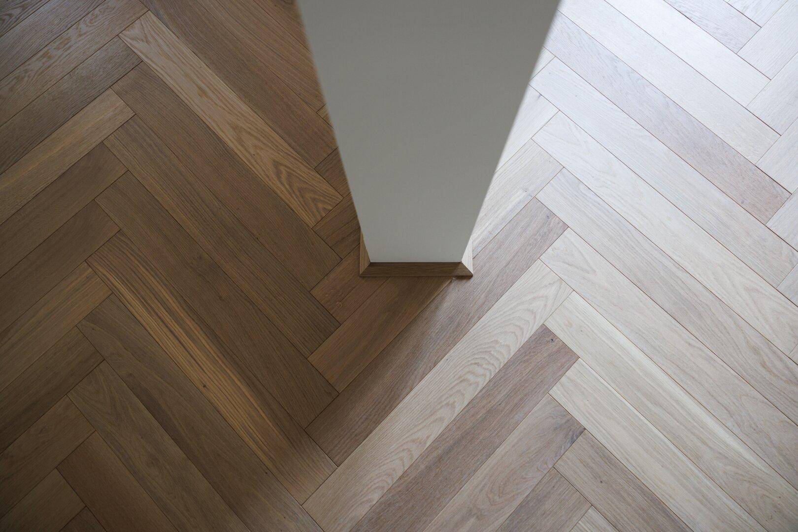 Detailaufnahme von Fischgrätparkett mit Farbnuancen und einem weißen Pfeiler im Raum.