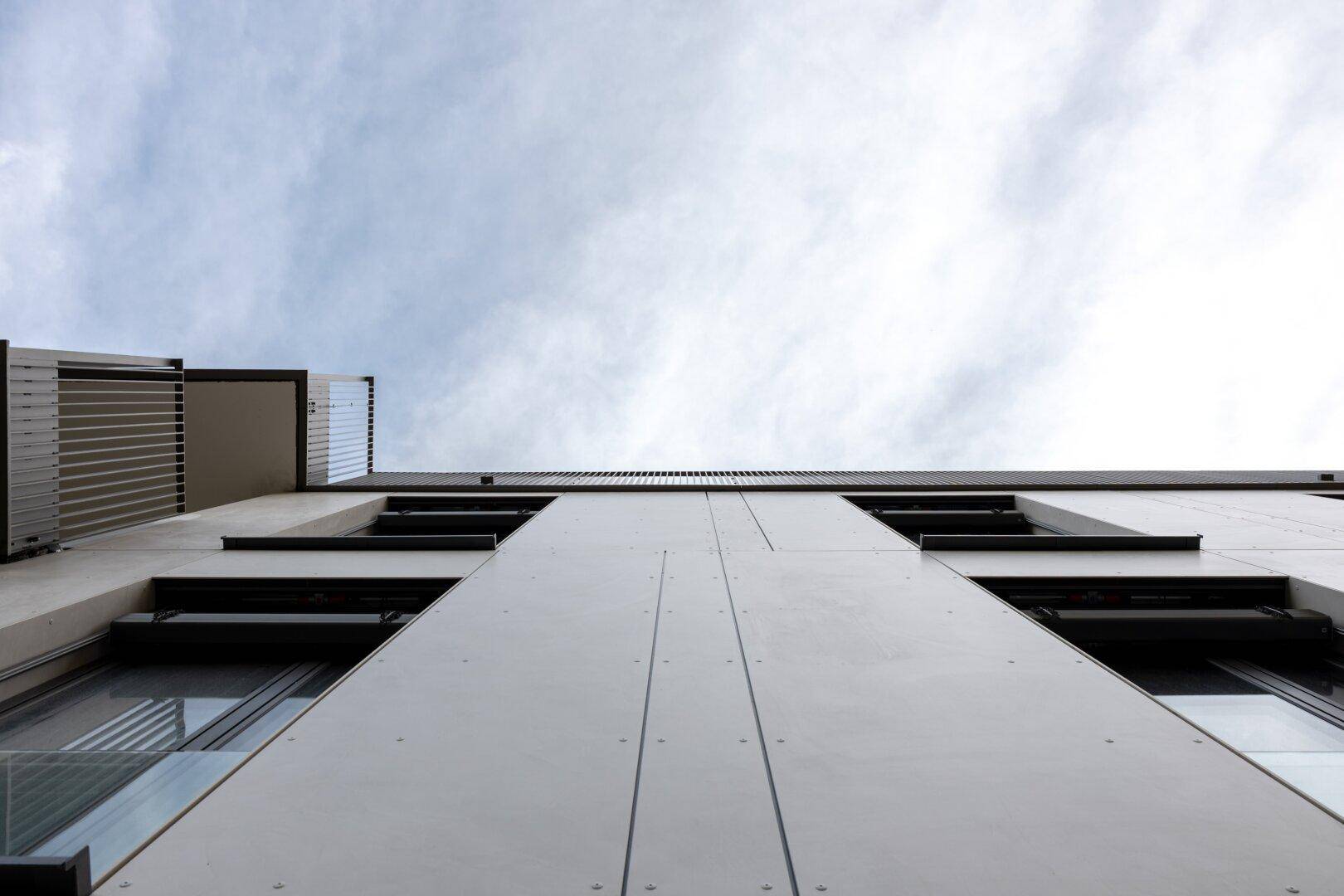 Blick nach oben auf eine moderne Gebäudefassade mit Balkonen und Fenstern unter bewölktem Himmel.