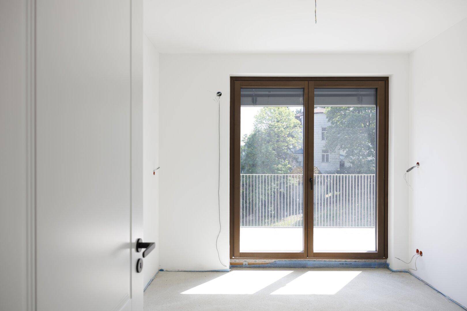 Unmöblierter Raum mit großen Fenstern, die viel Tageslicht hereinlassen und Zugang zu einem Balkon bieten.