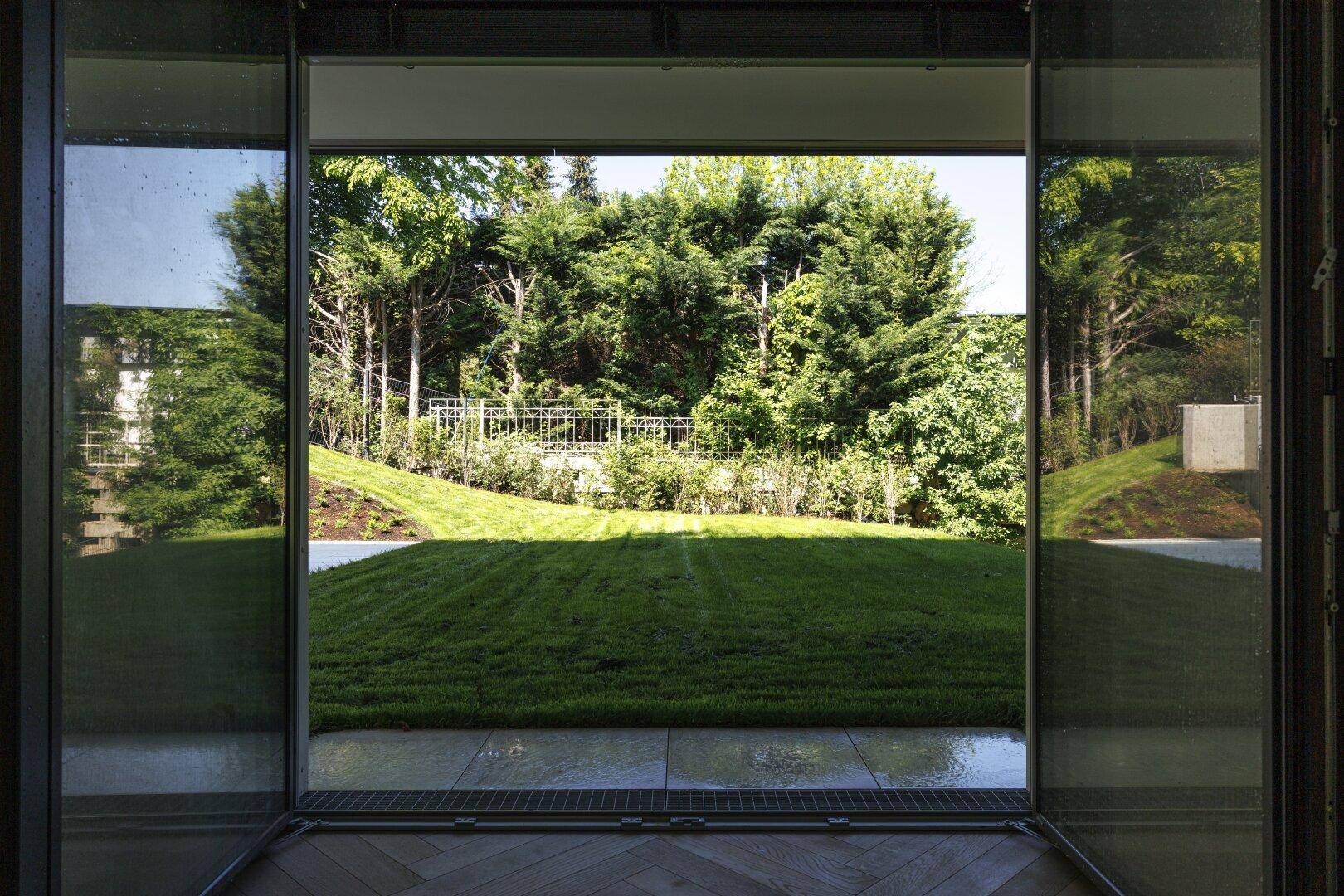 Blick aus einem Wohnraum durch große Glasfront auf einen gepflegten Garten mit Bäumen und Rasenfläche.
