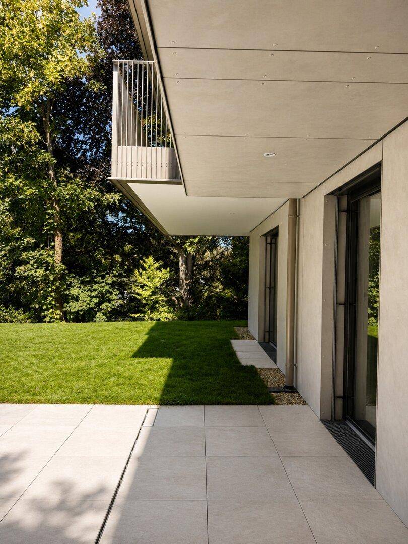Moderne Gebäudefassade mit großzügiger Terrasse, Rasenfläche und altem Baumbestand unter blauem Himmel.