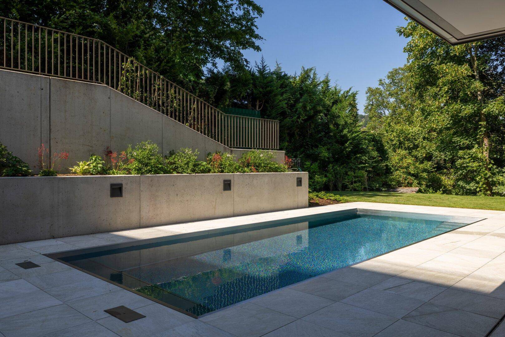 Weitläufiger Außenbereich mit rechteckigem Pool, umgeben von einer Betonmauer und viel Grün.