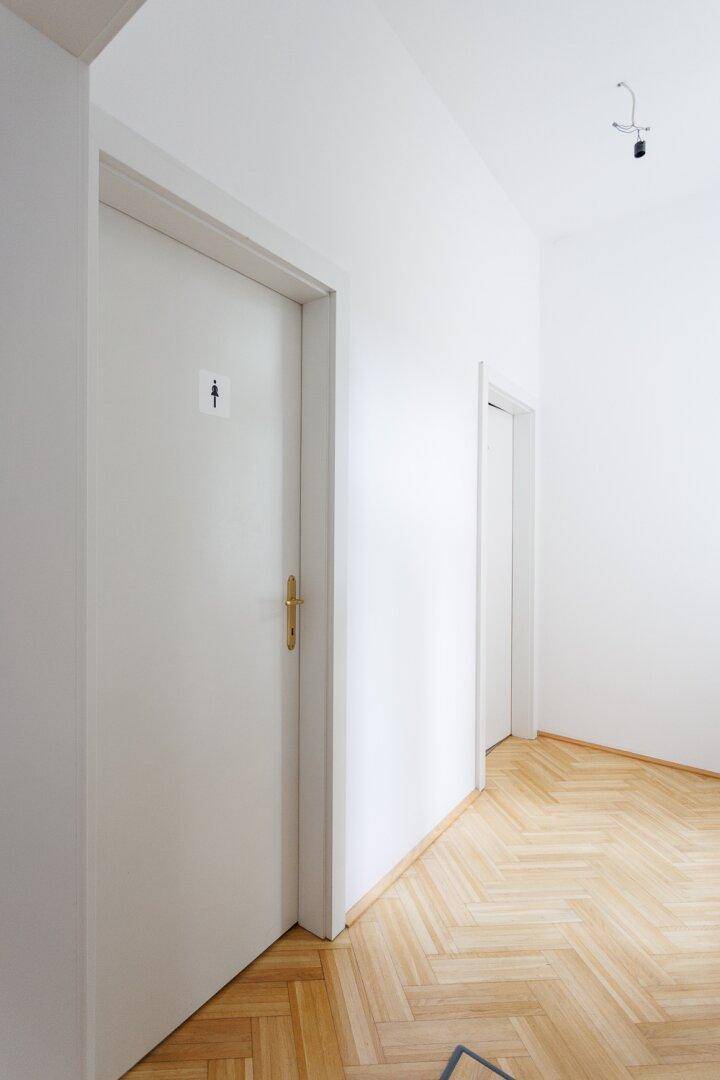 Weiße Tür zu einer Toilette mit Fischgrätparkett in einem hellen Flur.