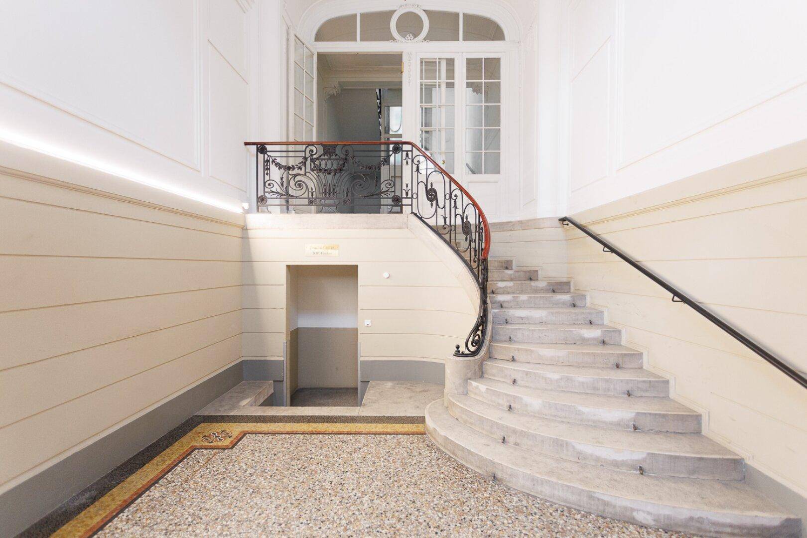 Elegantes Treppenhaus mit geschwungenem Geländer, Terrazzoboden und hohen Decken.