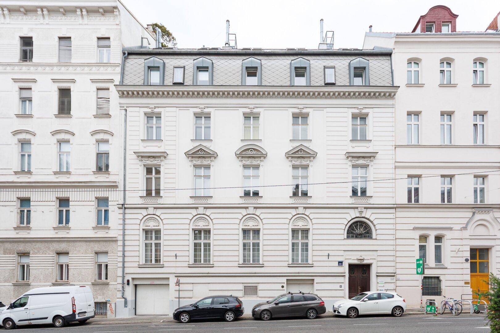Klassische Fassade eines Mehrfamilienhauses an einer belebten Straße mit parkenden Autos.