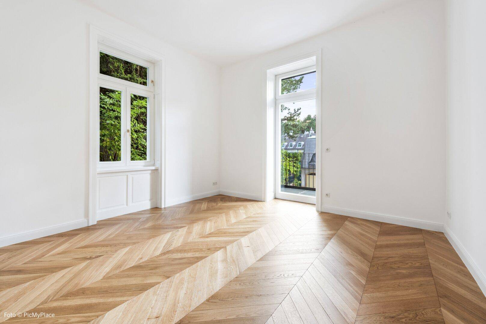 Helles Zimmer mit Fischgrätparkett, großem Fenster und Zugang zu einem französischen Balkon.