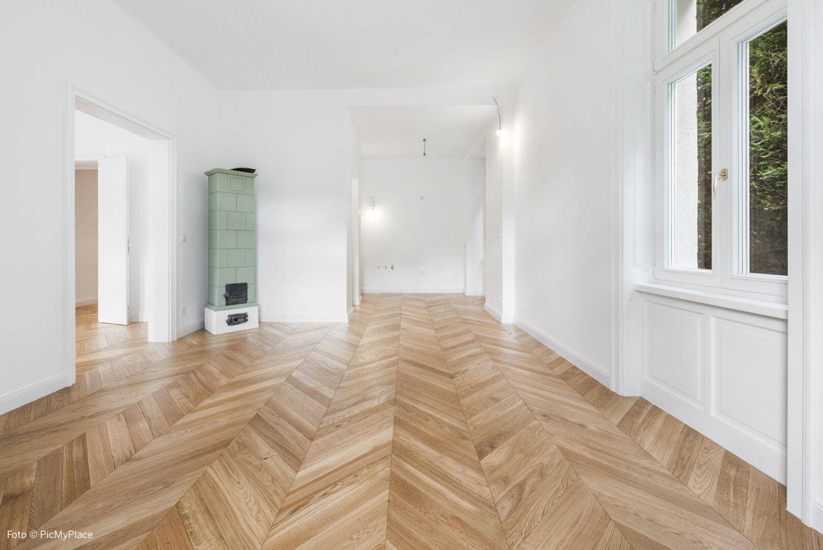 Helles Zimmer mit Fischgrätparkett und einem traditionellen grünen Kachelofen als Blickfang.