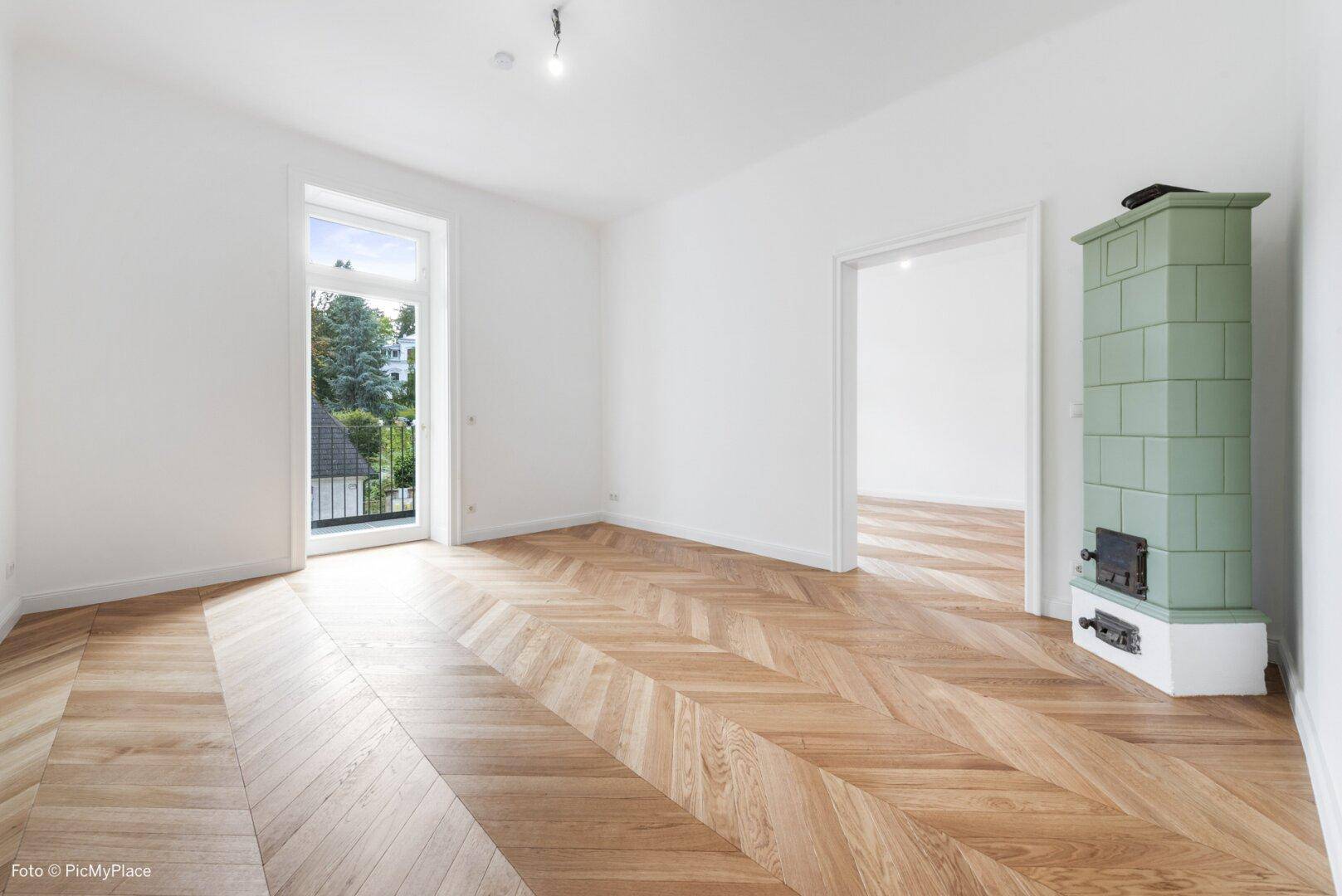 Geräumiges Zimmer mit Fischgrätparkett, Kachelofen und Zugang zu einem französischen Balkon.
