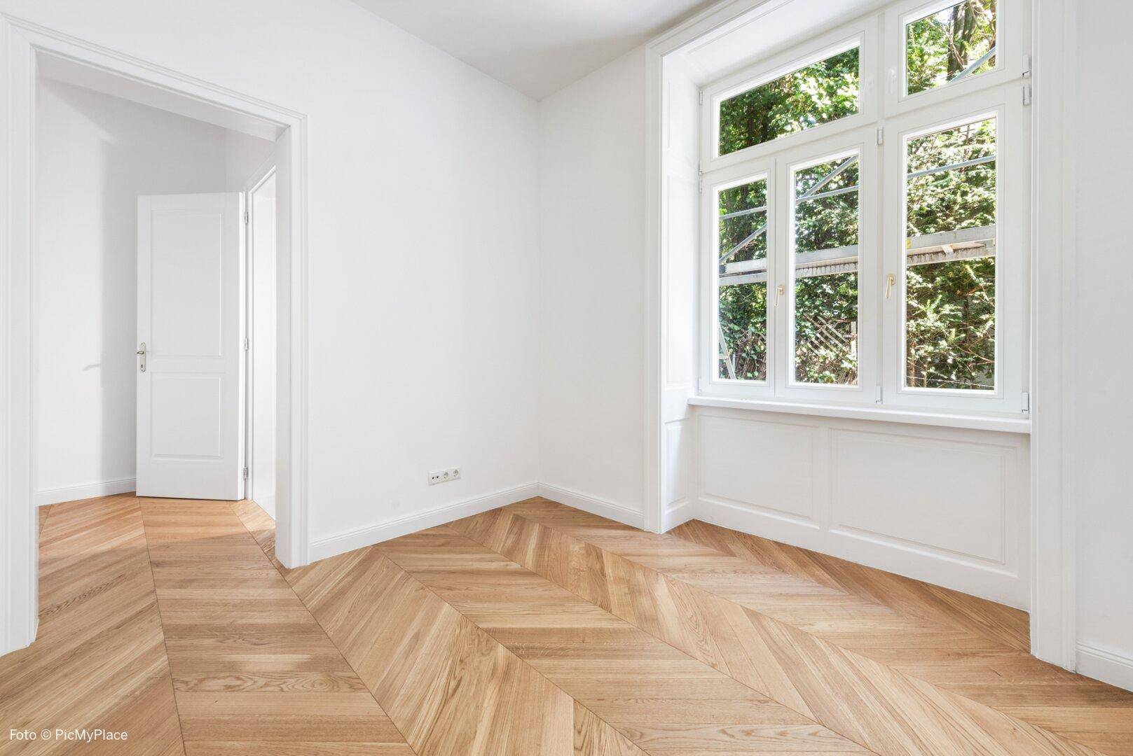 Geräumiges Zimmer mit Fischgrätparkett und einem großen Fenster mit klassischer Verkleidung.