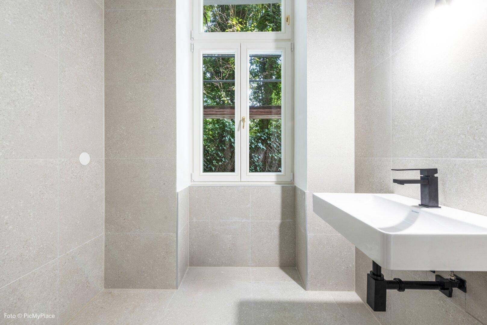 Modernes Badezimmer mit hellgrauen Fliesen, elegantem Waschbecken und Fenster für Tageslicht.