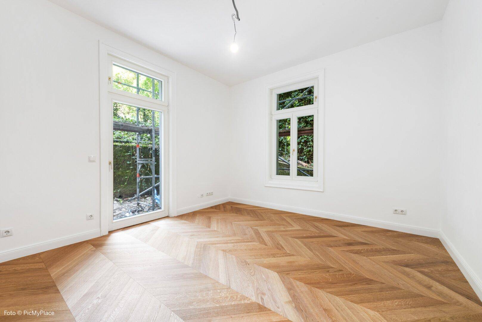 Helles Zimmer mit Fischgrätparkett, großem Fenster und Zugang zu einem französischen Balkon.
