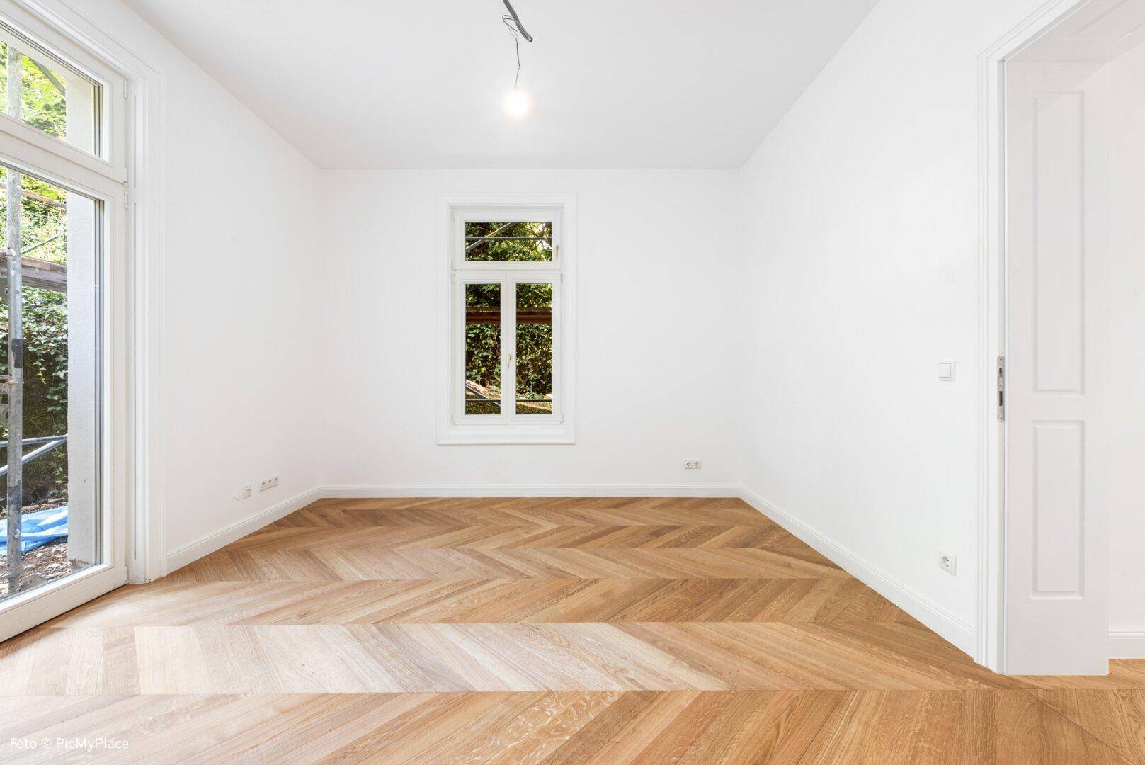 Helles Zimmer mit Fischgrätparkett, einem Fenster und Zugang zu einem französischen Balkon.