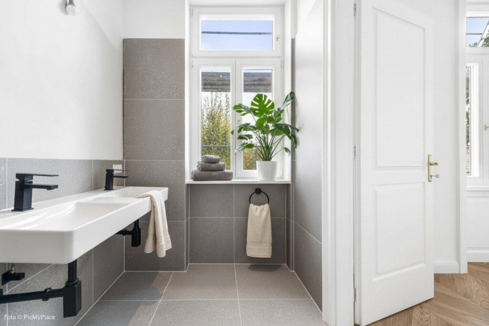 Modernes Badezimmer mit Doppelwaschbecken, grauen Fliesen und einem Fenster für Tageslicht.
